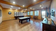 Located at 6950 Birdseye Ave NE in Olympia, Washington, this single-floor recreational facility is thoughtfully designed as a dedicated entertainment and game room space. The main area features warm golden-yellow walls, rich hardwood-style flooring, and a coffered ceiling with recessed lighting and stylish pendant fixtures that illuminate the room with a welcoming glow.

The billiards area is a focal point of the space, showcasing a full-size pool table centered beneath a striking multi-arm pendant light. A shuffleboard table lines the adjacent wall, accompanied by a curated gallery of billiard ball-themed framed artwork. Bar-height seating is positioned near windows fitted with blinds, allowing natural light to filter in. A door provides direct access to an outdoor area, further enhancing the open feel.

A separate card room is accessible via a glass-paneled door, offering a more intimate setting. This enclosed space features two round poker tables surrounded by cushioned leather chairs and is decorated with framed playing card artwork. Tufted bench seating lines the walls, creating a lounge-like atmosphere. Interior windows connect this room visually to the main space, maintaining a sense of openness while providing a quieter setting for card games. Together, these areas form a comprehensive and well-appointed recreational destination.