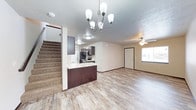 This two-story space features an open-concept main level that seamlessly connects the kitchen and living area. The kitchen is equipped with dark espresso-toned cabinetry, a peninsula with a light-colored countertop, and stainless appliances including a range and refrigerator. An overhead flush-mount light fixture illuminates the kitchen, while a stylish multi-bulb chandelier anchors the central dining or transitional area.

The living area extends naturally from the kitchen, finished with wood-look plank flooring that runs throughout the main level, lending warmth and visual continuity. A ceiling fan with a window nearby allows for natural light and ventilation. The neutral wall tones throughout provide a clean, versatile backdrop.

A carpeted staircase along one wall leads to the second floor, offering a clear visual separation between living levels. The overall layout is practical and functional, with an efficient flow between spaces. The combination of light walls, wood-look flooring, and dark kitchen cabinetry creates a balanced, contemporary aesthetic suited to everyday living.