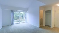 This two-floor space offers a clean, neutral interior designed for comfortable living. The upper level showcases a spacious, light-filled living area with vaulted ceilings that add a sense of openness and volume. Large sliding glass doors lead to a private balcony overlooking lush, tree-lined greenery, allowing abundant natural light to fill the room. Soft, neutral carpeting extends throughout the living area, contributing to a warm and inviting ambiance. A ceiling fan provides additional comfort overhead.

Adjacent to the living area, a dedicated laundry space with an in-unit washer is conveniently accessible behind a closet door. The lower level features a generously sized carpeted room, which opens to a bathroom equipped with warm wood-toned cabinetry and a vanity, visible through a connecting doorway.

The kitchen is efficiently laid out with wood-trimmed cabinetry, light countertops, a stainless steel sink, a dishwasher, and a full-size refrigerator. Light tile flooring and upper wall-mounted cabinets maximize storage and practicality. Throughout, the space maintains a consistent neutral palette, offering a versatile and move-in-ready environment.