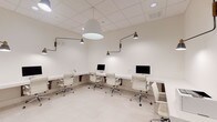 This single-floor workspace presents a sleek, minimalist aesthetic defined by an all-white palette and thoughtful design details. The room is configured with continuous built-in white desks arranged in an L-shaped layout along the walls, accommodating multiple workstations equipped with Apple iMac computers and contemporary white mesh rolling chairs with polished chrome frames.

Lighting is a standout feature of the space, combining a large dome-shaped pendant light at the center with an array of articulating wall-mounted arm lamps finished in matte black and brass. These directional fixtures provide focused task lighting across each workstation, contributing both function and a refined industrial-modern character to the otherwise understated interior.

The flooring consists of large-format light-toned tiles that reinforce the clean, open feel of the room. A drop ceiling with recessed grid panels and integrated recessed lighting further enhances the bright, evenly lit atmosphere. A frosted glass sliding door visible in one section of the space adds a degree of privacy and separation while maintaining visual connectivity to adjacent areas.

The overall ambiance is professional and polished, designed to support focused work in a shared environment. Every element — from the cohesive white surfaces to the carefully chosen fixtures — reflects a deliberate, design-forward approach to a functional workspace.