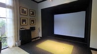 This single-floor space is purpose-built as an indoor golf simulator room, offering a focused and immersive environment for golf practice and entertainment. The centerpiece of the room is a large, wide-format projection screen framed by a bold black surround, creating a striking visual contrast against the neutral gray walls. A synthetic turf hitting mat in vibrant yellow-green is positioned directly in front of the screen, providing a realistic surface for drives and iron shots, with golf balls ready for play.

The room is thoughtfully appointed with golf-themed framed artwork arranged in a grid pattern on one wall, adding a refined, gallery-like quality to the space. Track lighting mounted on the ceiling delivers focused illumination directly onto the hitting area, ensuring optimal visibility during play. A sleek dark storage cabinet lines one wall, offering organized space for equipment and accessories, while a golf club stand keeps clubs neatly accessible.

The overall ambiance is intimate and purpose-driven, blending functional sports technology with tasteful interior design. The dark tones, clean lines, and curated décor give the room a polished, upscale feel — making it equally suited for serious practice sessions and casual entertainment.