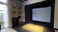 This single-floor space is purpose-built as an indoor golf simulator room, offering a focused and immersive environment for golf practice and entertainment. The centerpiece of the room is a large, wide-format projection screen framed by a bold black surround, creating a striking visual contrast against the neutral gray walls. A synthetic turf hitting mat in vibrant yellow-green is positioned directly in front of the screen, providing a realistic surface for drives and iron shots, with golf balls ready for play.

The room is thoughtfully appointed with golf-themed framed artwork arranged in a grid pattern on one wall, adding a refined, gallery-like quality to the space. Track lighting mounted on the ceiling delivers focused illumination directly onto the hitting area, ensuring optimal visibility during play. A sleek dark storage cabinet lines one wall, offering organized space for equipment and accessories, while a golf club stand keeps clubs neatly accessible.

The overall ambiance is intimate and purpose-driven, blending functional sports technology with tasteful interior design. The dark tones, clean lines, and curated décor give the room a polished, upscale feel — making it equally suited for serious practice sessions and casual entertainment.