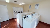This shared laundry facility is designed for practicality and ease of use. The single-floor space is outfitted with multiple top-loading washing machines arranged in a row along a central dividing counter, providing ample capacity for residents. Along the back wall, a bank of stacked dryer units maximizes the use of vertical space, further increasing the facility's overall laundry capacity.

A long, white folding counter runs through the center of the room, offering a dedicated surface for sorting and folding laundry. The walls are painted in a clean, neutral cream tone, contributing to a bright and sanitary atmosphere. Overhead fluorescent lighting ensures the room is well-lit throughout.

Instructional posters for both washing and drying are mounted on the walls, making the facility user-friendly for all occupants. The flooring features wood-look tile in a warm reddish-brown tone, adding a subtle visual warmth to the utilitarian space. Natural light filters in from one side of the room, complementing the overhead lighting. Overall, the facility is clean, organized, and efficiently laid out.