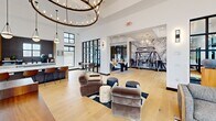 This expansive 2,590 square foot community space showcases a blend of modern design and functional amenities. The entrance features a spacious lobby with glass doors, providing a welcoming atmosphere. The fitness center is well-equipped with state-of-the-art cardio and strength training equipment, including treadmills, weight machines, and free weights. Large windows in the gym offer natural light and views of the outdoors.

The lounge areas are thoughtfully designed with a mix of contemporary and comfortable furnishings. A striking circular chandelier serves as a focal point in one room, while another area features a fireplace surrounded by cozy seating. The space incorporates a variety of seating options, from plush armchairs to bar-style seating, catering to different preferences and activities.

Hardwood flooring runs throughout, lending warmth to the contemporary design. Black-framed windows and glass partitions create a sense of openness while delineating different functional areas. Artistic elements, such as a large monochrome mural, add visual interest. The overall aesthetic is clean and inviting, with a neutral color palette accented by carefully chosen decorative elements, making it an ideal space for residents to gather, work, or relax.