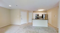 This single-floor unit features an open-concept layout that connects the kitchen and living area into one cohesive, light-filled space. The walls are finished in a soft, neutral tone that enhances the sense of brightness throughout.

The kitchen is outfitted with stainless steel appliances, including a refrigerator, electric range, and built-in microwave. White cabinetry extends to the upper walls, providing generous storage, while a peninsula with a dark countertop serves as both a functional workspace and a natural divider between the kitchen and living area. A sink is integrated into the peninsula, adding to the kitchen's practicality.

The living area features wall-to-wall carpeting in a light, neutral tone, contributing to a warm and comfortable ambiance. Two white interior doors are visible near the entryway — one serving as the main entrance and the other likely leading to additional rooms or a closet. Flush-mounted ceiling lighting provides consistent illumination throughout the space. The overall layout is clean, functional, and well-suited for everyday living.