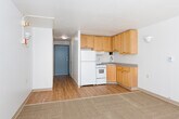 This single-floor unit offers a clean, functional layout filled with natural light and practical design. The main living area is generously proportioned, featuring a combination of hardwood-style flooring and neutral carpet, with crisp white walls and a textured ceiling. Large sliding glass doors dominate one wall, opening onto a private balcony that overlooks surrounding greenery and neighboring buildings, allowing abundant sunlight to pour into the space throughout the day. Wall-mounted light fixtures provide supplementary lighting and add a modest decorative touch to the living area. A sliding closet with floor-to-ceiling panels provides convenient built-in storage. The kitchen is efficiently arranged with warm oak cabinetry spanning both upper and lower sections, offering ample storage. It is equipped with a white refrigerator, an electric coil range, a built-in range hood, and a stainless steel sink set into a laminate countertop — all the essentials for comfortable day-to-day use. Hardwood-style flooring continues through the kitchen, maintaining visual continuity with the rest of the unit. The balcony, accessed via the sliding doors, features a metal railing and receives generous sunlight, making it a pleasant outdoor extension of the living space. Overall, the unit presents a straightforward, well-lit living environment with functional finishes throughout.