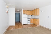 This single-floor unit offers a clean, functional layout filled with natural light and practical design. The main living area is generously proportioned, featuring a combination of hardwood-style flooring and neutral carpet, with crisp white walls and a textured ceiling. Large sliding glass doors dominate one wall, opening onto a private balcony that overlooks surrounding greenery and neighboring buildings, allowing abundant sunlight to pour into the space throughout the day. Wall-mounted light fixtures provide supplementary lighting and add a modest decorative touch to the living area. A sliding closet with floor-to-ceiling panels provides convenient built-in storage. The kitchen is efficiently arranged with warm oak cabinetry spanning both upper and lower sections, offering ample storage. It is equipped with a white refrigerator, an electric coil range, a built-in range hood, and a stainless steel sink set into a laminate countertop — all the essentials for comfortable day-to-day use. Hardwood-style flooring continues through the kitchen, maintaining visual continuity with the rest of the unit. The balcony, accessed via the sliding doors, features a metal railing and receives generous sunlight, making it a pleasant outdoor extension of the living space. Overall, the unit presents a straightforward, well-lit living environment with functional finishes throughout.