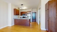 This single-floor residence showcases a cohesive design defined by warm bamboo hardwood flooring that flows seamlessly throughout every room, creating a bright and inviting atmosphere. The kitchen is a functional highlight, featuring rich cherry wood cabinetry, granite countertops, and a full suite of stainless steel appliances including a refrigerator, microwave, and stove. A peninsula with a built-in sink and pendant lighting above adds both style and practicality, while a wine rack integrated into the upper cabinetry adds a refined touch. The entry door is a distinctive wood-framed design with a frosted glass panel, lending a modern yet classic character to the space. The open floor plan, as seen in the dollhouse view, connects the kitchen to generously sized living and bedroom areas, offering a flexible layout. The bedroom is bright and airy, benefiting from two large windows with horizontal blinds that frame urban skyline views. The bathroom is well-finished with a granite-topped vanity, matching cherry cabinetry, a walk-in shower with glass enclosure, and matte black fixtures that complement the overall design palette. Clean white walls and white trim throughout contribute to a crisp, contemporary ambiance.
