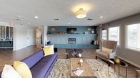 Lake Castleton's community amenity space is a thoughtfully designed single-floor gathering area that blends modern aesthetics with comfortable, functional living. The lounge area serves as the heart of the space, anchored by a striking teal accent wall with built-in wainscoting, a sleek electric fireplace, and a wall-mounted television — creating an inviting focal point ideal for socializing or relaxing. Plush seating, including a bold navy sofa with yellow accent pillows, adds a vibrant, contemporary touch complemented by warm wood-look flooring throughout.

The kitchen is a standout feature, showcasing a large waterfall-edge quartz island with an integrated sink and bar seating for multiple guests. Stylish pendant lighting hangs above, while floor-to-ceiling gray cabinetry, a subway tile backsplash, and stainless steel appliances — including a full-size refrigerator and microwave — round out the modern culinary setup.

Adjacent dining and conference areas are separated by sleek black-framed glass partitions, providing both visual openness and functional division of space. Edison-style pendant fixtures add an industrial-chic ambiance to these collaborative zones. A row of expansive windows in the lounge area floods the space with natural light and frames views of the outdoor pool area, further enhancing the bright and welcoming atmosphere throughout.