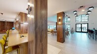 This expansive single-floor space is thoughtfully designed to accommodate a variety of social and functional needs within a cohesive, contemporary aesthetic. The entrance foyer sets the tone with large-format light stone tile flooring, curated wall art, and globe pendant lighting, leading visitors into the broader communal areas beyond.

Distinctive fluted wood columns serve as a recurring architectural element throughout, adding warmth and visual rhythm while defining separate zones without the need for full walls. The lounge area is anchored by generous cream sectional sofas layered with colorful throw pillows, vibrant abstract area rugs, and a marble-top coffee table, creating an inviting, residential feel. A wood-paneled media wall with a mounted television anchors one end of the seating area.

The bar and café zone commands attention with soaring double-height ceilings, dramatic textured charcoal accent walls, dark marble countertops, and an array of globe and dome pendant lights overhead. High-top tables and bar seating complement the space's lively atmosphere.

A co-working and dining area features long communal tables with bright yellow seating under linear pendant fixtures, alongside built-in shelving and beverage refrigeration. Floor-to-ceiling windows in a separate dining nook draw in natural light and offer outdoor garden views, completing this well-appointed, multifunctional environment.