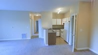 This single-floor residence offers a practical and functional layout designed for comfortable everyday living. The open-concept design connects the kitchen and living area seamlessly, creating a spacious and accessible flow throughout the main living space. The kitchen is equipped with white appliances, including a full-size refrigerator and electric range, complemented by a mix of white upper cabinetry and warm wood-toned lower cabinets. Dark countertops provide a subtle contrast, while a peninsula island with an integrated sink adds additional prep space and serves as a natural divider between the kitchen and the carpeted living area. Track lighting above the kitchen provides focused illumination. The living area features plush wall-to-wall carpeting, contributing to a cozy atmosphere. A hallway extends from the main living space, leading to additional rooms beyond, suggesting a well-organized bedroom and bathroom layout. The unit also includes a floor vent for climate control. Overall, the space presents a clean, neutral palette with beige walls and light-toned finishes, making it a versatile and move-in-ready environment suitable for a variety of living arrangements.