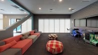 This contemporary single-floor amenity space is designed for socializing and recreation, combining distinct zones that flow cohesively together. A dramatic large cut-out opening connects a sunken lounge area to the main game room, framed by teal accent trim that serves as a bold architectural feature throughout. The lounge is anchored by a sprawling modular sectional in coral, red, and gray tones, complemented by plaid and checkered ottomans atop dark carpeting. Roller shades diffuse natural light along a full window wall, while an arcade game station adds entertainment nearby.

The adjacent game room features a shuffleboard table and pool table set against a patterned geometric accent wall, with interior windows offering sightlines between zones. A meeting and dining area boasts a vibrant multi-toned striped floor, a long communal table surrounded by turquoise chairs, an artistic Edison-bulb chandelier, and a mounted flat-screen TV. A high glass-block window brightens the space from above. Double doors lead to an outdoor terrace, and a ping-pong table anchors the open central floor. Together, these areas create a dynamic, well-appointed gathering environment.