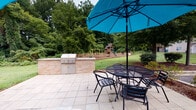Located at 609 Easy St in Kannapolis, North Carolina, this single-story property combines classic brick construction with practical outdoor amenities set against a backdrop of mature trees and open green space. The exterior showcases rich, multi-toned brick detailing alongside vinyl siding, giving the building a durable and timeless appearance. A covered concrete walkway runs along the building's length, supported by a warm wood-paneled ceiling and providing sheltered access along the brick facade. Entry steps are constructed of solid wood, adding a natural touch to the approach. The outdoor living space is a standout feature, highlighted by a spacious tiled patio complete with a built-in brick outdoor kitchen and grill station. A wrought iron dining set shaded by a vibrant teal umbrella offers a comfortable spot for outdoor gatherings. The expansive backyard is bordered by a white vinyl privacy fence on one side, with open views toward wooded surroundings on the other, creating a sense of seclusion and natural beauty. A children's wooden playground structure adds a family-friendly element to the grounds. Lush landscaping with mulched beds, mature trees, and well-maintained lawn areas complete the outdoor environment, making this property well-suited for both relaxation and entertaining.