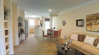 This single-floor residence offers a thoughtfully designed open-concept layout that seamlessly connects the main living areas, creating a spacious and cohesive environment. The living room provides a comfortable gathering space, anchored by a sleek dark coffee table and a plush sofa, with decorative accents adding a touch of personality to the neutral walls adorned with crown molding. Adjacent to the living room, a defined dining area features a round dark-topped table surrounded by upholstered chairs in warm terracotta tones, ideal for both everyday meals and entertaining guests. Crown molding runs throughout, lending a refined architectural detail to the interiors. The open kitchen is equipped with light wood cabinetry, a stainless steel refrigerator, and ample counter space. A raised breakfast bar serves as a natural divider between the kitchen and dining area, offering additional seating and casual dining options. Windows throughout the space invite natural light, brightening the warm beige walls and neutral carpeted flooring. The overall ambiance is warm, welcoming, and move-in ready, with a classic residential aesthetic suited for comfortable everyday living.