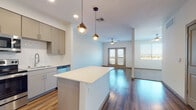 This contemporary single-floor residence showcases a thoughtfully designed open-concept layout that seamlessly connects the kitchen, dining, and living areas. The kitchen is outfitted with a full suite of stainless steel appliances, including a refrigerator, range, microwave, and dishwasher, complemented by taupe flat-front cabinetry, a clean white quartz countertop, and a central island ideal for meal prep or casual dining. Decorative globe pendant lights hang above the island, adding a warm, modern touch alongside recessed ceiling lighting throughout.

Rich hardwood-style flooring extends across the main living areas, creating a cohesive and inviting aesthetic. The living space is bright and airy, anchored by a ceiling fan and French doors that allow natural light to flow in while offering access to the exterior. A carpeted bedroom provides a quiet retreat, featuring a large window with outdoor views and a ceiling fan for comfort.

The bathroom is finished with a sleek white vanity featuring quartz countertops, an undermount sink with brushed nickel fixtures, and a backlit mirror that adds a sophisticated, modern flair. A tub-shower combination completes the bath. Throughout the space, clean white walls, consistent finishes, and well-placed lighting create a polished and functional living environment.