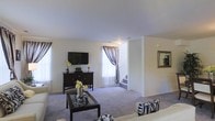 Located at 700 Lisa Place in North Brunswick, NJ, this two-story home offers a welcoming and well-appointed layout across two levels. The main floor features a spacious living area finished with plush carpeting, creating a comfortable and cozy atmosphere. Large windows dressed with elegant striped curtains allow generous natural light to fill the room, enhancing the bright and airy feel of the space. A media console with wall-mounted television serves as a central focal point, complemented by decorative accents and floral arrangements that add warmth to the interior. A stylish coffee table anchors the seating area, which flows seamlessly into the rest of the main level. A centrally positioned staircase with light-colored risers connects the main floor to the upper level, providing both functional access and a natural visual transition between floors. The neutral color palette throughout — from the white walls and ceilings to the soft gray carpeting — gives the home a clean, versatile aesthetic that accommodates a variety of décor styles. Overall, the space presents a comfortable, move-in-ready home with classic design elements and a practical two-floor layout suited for everyday living.