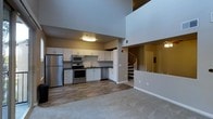 This two-story space makes an immediate impression with its dramatic double-height ceilings, which lend an open and airy atmosphere to the main living area. The ground floor seamlessly integrates the kitchen and living space, with a clear distinction in flooring — warm hardwood-style planks define the kitchen zone, while plush carpeting anchors the living area.

The kitchen is well-appointed with a full suite of stainless steel appliances, including a refrigerator, electric range, built-in microwave, and dishwasher. Crisp white cabinetry lines the walls, providing generous storage, and is complemented by a clean countertop workspace and a single-basin sink. The overall design is modern and functional, offering everything needed for everyday living.

A standout architectural feature is the elegant spiral staircase, which rises gracefully from the main floor to the upper level. The upper floor overlooks the living area below through an open railing, reinforcing the sense of vertical space and connectivity between the two levels. The upper floor also provides additional living or utility space, accessible via a doorway visible from below.

Overhead lighting on the main floor ensures the space is well-lit, and the tall walls contribute to the expansive, open feel that defines this thoughtfully designed two-level layout.