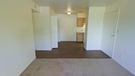 This single-floor space offers a straightforward, functional layout designed for comfortable everyday living. The living area is finished with soft carpeting underfoot, creating a warm and inviting atmosphere, while the adjoining open space transitions to wood-style hard flooring that extends toward the kitchen. Bright white walls throughout lend the interior a clean, neutral aesthetic that provides a versatile backdrop for a variety of décor styles. The open-plan arrangement between the living and kitchen areas promotes a sense of connected, flowing space. The kitchen is equipped with warm-toned wooden cabinetry, a white range, and a refrigerator, offering practical storage and cooking functionality in a compact footprint. A flush-mount ceiling light provides ambient illumination to the central area, and overhead vents are thoughtfully positioned for climate comfort. Structural dividing walls help define the separate zones within the space without sacrificing the overall openness. The unit's simple, uncluttered design makes it easy to personalize, while the combination of carpet and hard flooring adds both texture and practicality to the living environment.