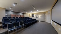 This private screening room delivers a true cinema experience within a compact, well-designed footprint. The space is arranged with tiered, stadium-style seating rows that ensure unobstructed sightlines from every position. Seating consists of black leather-style reclining chairs with integrated cup holders, accompanied by additional row-style chairs along the front, accommodating a generous number of viewers in comfort. At the front of the room, a large projection screen dominates the wall, framed cleanly against the neutral warm-toned walls and supported by in-wall surround sound speakers positioned throughout the space for an immersive audio experience. A ceiling-mounted projector provides the image source. The flooring features a durable patterned carpet, and step lighting along the tiered aisles ensures safe navigation in low-light conditions. Wall sconces provide ambient lighting, complementing the recessed ceiling lights that illuminate the room when not in screening mode. Framed movie posters adorn the walls, reinforcing the cinematic atmosphere. At the rear of the room, shelving displays an extensive collection of physical media. Multiple entry and exit doors provide convenient access. The overall design is functional, comfortable, and purpose-built for enjoying films in a communal yet private setting.