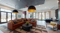 Located at 309 Mirroton Ave in Lakeland, Florida, this single-floor community clubhouse offers a thoughtfully designed gathering space that blends modern aesthetics with functional amenities. The main lounge area is anchored by a large cognac leather sectional sofa set atop a bold geometric-patterned area rug, complemented by dramatic oversized pendant lights with gold interiors and a dark ceiling treatment that adds depth and contrast. Vibrant contemporary artwork punctuates the walls, lending an artistic energy throughout the space. Large windows dressed with sheer curtains flood the interior with natural light and offer views of the surrounding landscape. The game room features a full-size billiards table, a wall-mounted flat-screen TV, and a bar-height counter with seating, all set against a striking textured dark accent wall. A well-appointed kitchen includes grey cabinetry with white quartz countertops, stainless steel appliances, a beverage refrigerator, and a bold white tile backsplash. An adjacent service area features a U-shaped counter layout with interior-facing sliding barn-style windows, ideal for events. A sleek seating nook near the entrance offers a welcoming reception area with modern swan chairs and curated décor. Wood-look tile flooring and recessed lighting complete the polished, contemporary feel.