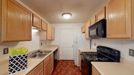 This single-floor residence offers a functional and straightforward layout across several well-defined spaces. The galley-style kitchen is equipped with light wood cabinetry providing ample storage above and below the counters, complemented by light-toned countertops, a double basin sink, and a full suite of black appliances including an electric range, built-in microwave, dishwasher, and refrigerator. The flooring features a warm parquet-style pattern, adding visual interest to the space.

The living area is carpeted and opens to a separate dining space, distinguished by hardwood-style flooring and a hanging chandelier. A sliding glass door in the living area is fitted with vertical blinds and offers views of surrounding greenery, allowing natural light to fill the room.

The bedrooms are carpeted and feature neutral walls with natural light provided by windows overlooking outdoor greenery. One bedroom includes a ceiling fan with light fixture. The two bathrooms each include a tub-shower combination, toilet, and vanity with storage cabinetry. One bathroom features a dark granite-style countertop, while the other offers a lighter wood-toned vanity. Both are finished with light ceramic tile flooring.