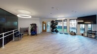 This single-floor fitness and wellness studio is thoughtfully designed to accommodate a variety of exercise and therapeutic activities. The main exercise area features light wood-look flooring throughout, providing a clean, cohesive aesthetic that extends across the entire space. Circular halo pendant lights are suspended from the white ceilings, delivering bright, even illumination across the open floor areas.

One side of the main workout zone is lined with floor-to-ceiling dark-tinted glass panels and a ballet-style barre, creating a dedicated movement or dance area with a reflective, studio-like atmosphere. Adjacent to this, a well-equipped equipment zone houses medicine balls, resistance bands, stability equipment, and various training accessories neatly organized on rolling carts and wall-mounted racks.

Large windows along one wall invite natural light and offer views of the surrounding outdoor area, enhancing the open and energizing ambiance. A separate section of the space transitions into a reception or waiting area, where soft blue-grey walls, seating chairs, and white paneled doors lead to private rooms suitable for consultations or treatment sessions. The overall design balances functional training space with a calm, professional environment.