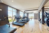 The Fitness Room at 1 Hanson St in Lynn, MA is a generously sized, well-appointed exercise facility spanning a single floor. The space is divided into two distinct functional zones, each thoughtfully designed to support a comprehensive workout experience.

The cardio zone features a decorative hardwood floor with colorful geometric inlays, lending the area a dynamic, modern aesthetic. A large row of treadmills lines one side of the room, accompanied by elliptical trainers and stationary bikes. Exposed industrial-style ductwork runs across the ceiling, adding an urban character to the space. Floor-to-ceiling windows and large glass garage-style doors flood the area with natural light and offer open views of the surrounding outdoor courtyard and greenery, creating an energizing atmosphere.

The strength training zone transitions to durable rubber tile flooring, providing a practical surface for heavy equipment. This area is equipped with a multi-functional cable machine, a power rack with barbells and weight plates, a full dumbbell rack, and a selection of selectorized strength machines. Large windows continue throughout this section, maintaining the bright and airy feel of the overall space.

Recessed lighting supplements the abundant natural light, and the neutral gray wall palette throughout the fitness room presents a clean, focused environment suitable for all types of workouts.