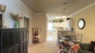 This single-story home offers an open-concept floor plan that seamlessly connects the living, dining, and kitchen areas into one cohesive, functional space. Warm beige walls are complemented by classic crown molding that runs throughout, lending the interior a polished, traditional character.

The kitchen is outfitted with white cabinetry that extends to the upper walls, providing generous storage space. White appliances, including a range and built-in microwave, are neatly integrated, while a pendant light fixture with a bell-shaped shade hangs above the counter, adding a decorative focal point. A window above the sink draws in natural light, brightening the space. Upper cabinet tops are styled with small potted plants and decorative accents.

The adjoining dining area accommodates a glass-top table surrounded by ornate wrought-iron chairs, creating an elegant yet casual setting for meals. A decorative ladder shelf nearby offers additional display space.

Neutral carpeting flows through the living areas, contributing to the home's warm, inviting ambiance. The layout is practical and well-proportioned, offering a comfortable, cohesive living environment throughout.