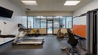 Located at 80 N Portage Path in Akron, Ohio, this fitness center provides a comprehensive workout facility within a compact, efficiently utilized single-floor space. The gym features a drop-panel ceiling with recessed fluorescent lighting that ensures consistent, even illumination throughout the room. Full-length mirrors line one wall, visually expanding the space and allowing users to monitor their form during exercise.

The room is outfitted with a diverse selection of equipment to support a full-body workout routine. Cardio options include multiple treadmills by Spirit and Landice, an elliptical trainer, a stationary air bike, and a recumbent bike. Strength training is well-represented with a Precor multi-functional cable machine, a multi-station weight system, free weights, and additional resistance equipment. An orange exercise mat is available for floor-based training.

A wall-mounted television and a clock keep users informed and entertained during workouts, while sanitation supplies are conveniently stationed nearby. Large glass panel windows and a door on one wall connect the gym visually to an adjacent indoor pool area, adding an open, communal feel to the space. The dark rubber flooring throughout provides durability and traction, making this fitness center a practical and fully functional amenity.