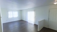 This single-floor residential space offers a clean, minimalist layout with a bright and open atmosphere. The main living area is defined by rich, dark hardwood flooring that contrasts elegantly with the crisp white walls and textured ceiling, creating a warm yet contemporary feel. Natural light floods the space through multiple large windows dressed with vertical blinds, including a prominent sliding glass door that opens to the exterior and further enhances the sense of openness. The white walls throughout contribute to a fresh, neutral palette that provides a versatile backdrop for any interior style. A partial half-wall partition adds a subtle architectural detail, gently delineating areas within the open floor plan without disrupting the flow of the space. The textured ceiling adds a layer of visual interest overhead, while clean white baseboards frame the flooring throughout. Overall, the space presents a straightforward, well-lit, and functional layout well-suited for comfortable everyday living.