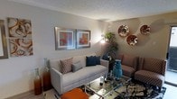 Located in Euless, TX, this single-story space offers a thoughtfully designed living area that balances comfort and style. The living room serves as the focal point of the space, featuring a light gray sofa accented with patterned and teal throw pillows, complemented by upholstered accent chairs in a rich geometric print. A sleek glass-top coffee table anchors the seating arrangement, reflecting light and contributing to an open, airy feel. Decorative touches include a pair of framed botanical artwork pieces in warm orange and red tones, alongside metallic wall décor that adds texture and visual interest to the neutral gray walls. A tall potted plant in the corner introduces a natural element, enhancing the overall ambiance. The space is illuminated by a stylish arc floor lamp, creating warm, focused lighting throughout the room. The textured ceiling adds architectural character, while the patterned area rug grounds the furniture arrangement. Overall, the space presents a cohesive, contemporary aesthetic with a warm color palette of teal, orange, and neutral tones that creates an inviting and well-appointed atmosphere.