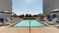 This outdoor amenity deck offers a well-appointed resort-style retreat designed for relaxation and leisure. The centerpiece is a generously sized rectangular swimming pool with clear, turquoise water, accessible via stainless steel handrails and surrounded by a wide concrete pool deck. Rows of cushioned chaise lounges and shaded umbrella seating are arranged throughout, providing ample space for sunbathing and unwinding. A separate spa with its own stainless steel entry rails is nestled nearby, framed by a sleek privacy wall and decorative planters. Comfortable oversized round daybeds, accented with vibrant blue, yellow, and teal throw pillows, offer an inviting lounging alternative. The deck also features thoughtfully designed landscaping, with mature olive trees and lush garden beds integrated into raised planters that add natural texture and greenery to the space. Glass railings along the perimeter preserve unobstructed views of the surrounding hillside and tree-lined horizon. The contemporary building architecture, characterized by clean lines, large windows, and private balconies, encloses portions of the deck to create a semi-private courtyard atmosphere. The space is bathed in abundant natural sunlight, enhancing its open, airy ambiance throughout the day.