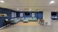 This single-floor space is designed as a stylish and comfortable lounge area, combining modern aesthetics with a welcoming ambiance. The focal point of the room is a striking navy blue accent wall, which creates a bold, sophisticated backdrop for the space. A large wall-mounted flat-screen TV is centrally positioned on this wall, flanked by a pair of framed geometric artwork pieces in coordinating blue tones, adding a cohesive and curated feel to the design.

The seating arrangement is generous and inviting, anchored by an expansive light blue sectional sofa that wraps around a neutral-toned area rug. Decorative throw pillows in navy and white patterns add texture and visual interest to the sectional. A matching ottoman serves as a functional centerpiece, while an accent chair in warm cognac provides a contrasting pop of color and additional seating.

The ceiling features a recessed tile grid with integrated lighting, providing bright, even illumination throughout the space. The flooring consists of large-format light grey tiles, contributing to a clean and contemporary look. Overall, the space exudes a relaxed yet polished atmosphere, well-suited for communal lounging, media viewing, or casual social interaction.