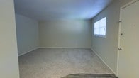 This single-story space presents a straightforward and functional layout, centered around a generously sized room finished with wall-to-wall carpeting in a neutral beige tone. The walls are painted in a soft, muted green-gray hue, contributing to a calm and understated atmosphere throughout. A window fitted with horizontal blinds is positioned along one wall, allowing natural light to filter into the room while maintaining privacy. The ceiling is smooth and painted white, complementing the clean lines of the space. White baseboards run along the perimeter of the room, providing a crisp contrast to the carpeted floor. Electrical outlets are evenly distributed along the walls, offering practical utility for a variety of uses. The transition from carpet to what appears to be a hard-surface flooring is visible at the room's edge, suggesting an adjoining area or entryway. Overall, the space is move-in ready, offering a blank canvas that is well-suited for a living area, bedroom, or flexible multi-purpose room. Its simple, uncluttered design makes it highly adaptable to a range of furnishing styles and functional needs.