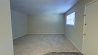 This single-story space presents a straightforward and functional layout, centered around a generously sized room finished with wall-to-wall carpeting in a neutral beige tone. The walls are painted in a soft, muted green-gray hue, contributing to a calm and understated atmosphere throughout. A window fitted with horizontal blinds is positioned along one wall, allowing natural light to filter into the room while maintaining privacy. The ceiling is smooth and painted white, complementing the clean lines of the space. White baseboards run along the perimeter of the room, providing a crisp contrast to the carpeted floor. Electrical outlets are evenly distributed along the walls, offering practical utility for a variety of uses. The transition from carpet to what appears to be a hard-surface flooring is visible at the room's edge, suggesting an adjoining area or entryway. Overall, the space is move-in ready, offering a blank canvas that is well-suited for a living area, bedroom, or flexible multi-purpose room. Its simple, uncluttered design makes it highly adaptable to a range of furnishing styles and functional needs.