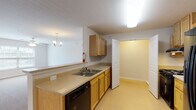 This single-floor unit offers a practical and accessible layout across several well-defined living spaces. The open living and dining area is generously sized, finished with neutral carpet and illuminated by a combination of a ceiling fan with integrated lighting and an elegant chandelier. A window provides natural light and an outward view, enhancing the room's airy feel.

The galley-style kitchen is efficiently designed, featuring light oak cabinetry with ample upper and lower storage, beige laminate countertops, a double-basin sink, a black electric range with overhead ventilation, a black dishwasher, and a stainless steel refrigerator. The kitchen connects seamlessly to a utility or pantry area via a panel door, adding functional storage. A pass-through counter opens toward the dining space, maintaining an open connection between the two areas.

Two bathrooms serve the unit. One includes a full tub and shower combo with grab bars, an oak vanity with a framed mirror, and warm lighting. The second features a roll-in style shower with grab bars and a matching vanity, underscoring the unit's accessibility-conscious design.

A spacious walk-in closet with wire shelving and carpet flooring provides substantial storage. Throughout the unit, light-toned walls and consistent finishes create a clean, cohesive ambiance.