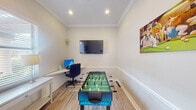 This single-room space serves a dual purpose as both a recreation and work area, thoughtfully designed to balance productivity with leisure. The room features light wood-look flooring and crisp white walls accented by classic crown molding and a white chair rail, lending a clean, polished aesthetic throughout.

A full-size foosball table anchors the center of the room, making it an immediate focal point for entertainment. A wall-mounted flat-screen television is positioned prominently on one wall, ideal for gaming or media viewing. Recessed ceiling lighting provides even, functional illumination across the space.

Along one wall, a white L-shaped desk houses a desktop computer setup with an ergonomic office chair, creating a capable workstation. A secondary side table offers additional surface space nearby. Natural light filters into the room through a window fitted with white horizontal blinds, adding brightness without sacrificing privacy.

Playful wall art featuring dogs in billiards scenes adds personality and a lighthearted atmosphere to the room. A glass-panel door provides access to an adjacent area. The overall design is compact yet efficiently arranged, making it a versatile space suited for both focused work and casual recreation.