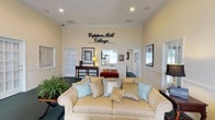 The Copper Mill Village Clubhouse is a thoughtfully designed single-story community facility that blends classic architectural details with a warm, welcoming atmosphere. The entrance features a striking arched transom window above a set of glass-paneled French doors, flooding the foyer with natural light and offering views of the surrounding brick exterior and landscaped grounds. Hardwood flooring at the entry transitions to rich teal carpeting throughout the interior, adding a cohesive and polished look to the space.

The main lounge area serves as the heart of the clubhouse, anchored by a cream-upholstered sofa adorned with soft blue and gold accent pillows, complemented by additional seating including a red chaise lounge. Elevated ceilings with recessed lighting and crisp crown molding lend an air of sophistication, while the prominent Copper Mill Village wall signage adds a strong sense of community identity.

Warm beige walls trimmed with classic chair-rail wainscoting carry throughout the space, creating a consistent and refined aesthetic. Decorative touches such as framed artwork, table lamps, and lush potted greenery enhance the inviting ambiance. Multiple doorways lead to adjacent rooms and corridors, suggesting additional functional spaces within the facility. Windows dressed with neutral drapery panels allow natural light to filter through while maintaining a comfortable and intimate environment.