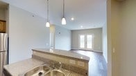 This well-designed single-floor residence offers a seamless open-concept layout that connects the kitchen and living areas, creating a spacious and cohesive environment. The kitchen is anchored by a generous granite countertop peninsula with a warm, speckled finish, complemented by a stainless steel double sink. Pendant lights hang gracefully from the ceiling above the peninsula, providing focused task lighting while adding a decorative touch. Recessed lighting throughout ensures the space remains bright and welcoming. The kitchen flows naturally into the living area, which features hardwood-style flooring that extends throughout, lending warmth and continuity to the space. Neutral, light gray walls create a clean and versatile backdrop. A standout feature of the living area is the set of classic French doors, framed in white and allowing natural light to pour in, enhancing the open and airy feel of the room. The vaulted ceiling adds a sense of height and grandeur, making the overall space feel larger and more expansive. The combination of modern finishes, thoughtful lighting, and architectural details such as the French doors and raised ceiling make this a functional yet stylish residential space suited for comfortable everyday living.