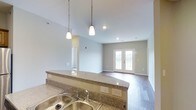 This well-designed single-floor residence offers a seamless open-concept layout that connects the kitchen and living areas, creating a spacious and cohesive environment. The kitchen is anchored by a generous granite countertop peninsula with a warm, speckled finish, complemented by a stainless steel double sink. Pendant lights hang gracefully from the ceiling above the peninsula, providing focused task lighting while adding a decorative touch. Recessed lighting throughout ensures the space remains bright and welcoming. The kitchen flows naturally into the living area, which features hardwood-style flooring that extends throughout, lending warmth and continuity to the space. Neutral, light gray walls create a clean and versatile backdrop. A standout feature of the living area is the set of classic French doors, framed in white and allowing natural light to pour in, enhancing the open and airy feel of the room. The vaulted ceiling adds a sense of height and grandeur, making the overall space feel larger and more expansive. The combination of modern finishes, thoughtful lighting, and architectural details such as the French doors and raised ceiling make this a functional yet stylish residential space suited for comfortable everyday living.