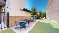 This urban outdoor space in Pensacola offers a versatile environment for recreation and relaxation. The area is characterized by a large expanse of artificial turf, providing a low-maintenance green space. A prominent feature is the tall brick wall that serves as a backdrop, adding a sense of privacy and urban charm. The space includes outdoor exercise equipment, encouraging physical activity. Comfortable seating areas with tables and chairs are available, perfect for socializing or outdoor dining. Landscaping elements, including trees and shrubs, soften the urban setting and provide natural shade. The area is partially enclosed by a metal fence, offering security while maintaining an open feel. Adjacent to the space is a building with a covered entrance, suggesting this could be a communal area for residents or visitors.