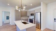 This contemporary single-floor residence offers a thoughtfully designed layout with clean finishes and an abundance of natural light throughout. The kitchen serves as the centerpiece of the space, showcasing flat-front cabinetry in a warm taupe finish with brushed gold hardware, a classic white subway tile backsplash, and a full suite of stainless steel appliances including a range, built-in microwave, and refrigerator. A generous kitchen island with a crisp white quartz countertop and an undermount sink anchors the space, while a striking brass and glass bubble chandelier adds a sculptural, modern touch overhead. Light oak hardwood flooring flows seamlessly throughout the unit, complementing the neutral white walls and creating a bright, cohesive aesthetic. The bedrooms are well-proportioned and filled with natural light from large windows that frame views of the surrounding neighborhood. A walk-in closet with dual hanging rods provides ample storage. The bathroom features a double vanity with white quartz countertops, undermount sinks, contemporary chrome fixtures, and a large wall mirror illuminated by overhead lighting. The overall ambiance is fresh, modern, and move-in ready.