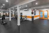 This fitness center at 300 W 2nd St in Santa Ana, California, offers a well-equipped, single-floor workout facility designed to support a comprehensive exercise routine. The space is defined by clean white walls accented with a bold orange horizontal stripe, dark rubber flooring for durability and safety, and recessed ceiling lighting that ensures bright, even illumination throughout.

The cardio zone is equipped with multiple treadmills and elliptical machines, each positioned for comfortable spacing. Multiple wall-mounted flat-screen TVs are placed strategically throughout the room, allowing users to stay entertained during their workouts. Structural columns are integrated seamlessly into the layout without disrupting the flow of the space.

A dedicated strength-training area features a multi-function cable and weight machine, providing options for a variety of resistance exercises. A free weights section includes a tiered dumbbell rack stocked with a range of weights, accompanied by an adjustable workout bench — ideal for targeted strength work. Medicine balls mounted on a wall rack add additional versatility for functional training.

The facility is finished with practical amenities including sanitation dispensers, a waste receptacle, and clear signage to support a clean and organized environment. Access is provided through a distinctive yellow-framed frosted glass door, adding a touch of design continuity to the overall aesthetic.