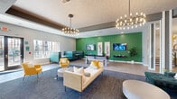 This generously sized single-floor amenity space is designed with a modern, vibrant aesthetic that blends functionality with comfort. A bold color palette—anchored by signature green accent walls, warm yellows, and teal tones—creates a cohesive and energetic atmosphere throughout. The main lounge area serves as the centerpiece, featuring plush seating arrangements, a tufted teal sofa, cream couches with yellow accent pillows, and a billiard table positioned beneath wall-mounted televisions. Statement multi-arm chandeliers illuminate the space, complementing the textured ceiling and open layout.

Adjacent to the lounge, a café-style dining nook offers round white tables and gold upholstered chairs beside large windows overlooking an outdoor courtyard and pergola area. The fully equipped kitchen features white shaker cabinetry, stainless steel appliances, subway tile backsplash, open shelving, and dark countertops—ideal for communal use.

A dedicated lobby area houses a bank of package lockers and a communal work table with colorful seating. Private study nooks with warm yellow walls and sconce lighting provide focused workspaces. Wood-look flooring and carpet zones delineate functional areas, while natural light flows in through multiple large windows.