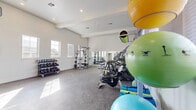 This fitness center is a spacious, single-floor facility designed to accommodate a comprehensive range of workout routines. The room features soaring ceilings with a coffered tray detail and recessed lighting, lending an open, airy quality to the space. Crisp white walls and large windows fitted with horizontal blinds allow generous amounts of natural light to fill the room, creating a clean and motivating environment.

The rubber-floored space is organized to support both cardio and strength training. Along one wall, a row of Matrix treadmills and elliptical trainers is positioned in front of a large mirror, maximizing the sense of depth and allowing users to monitor their form. A stationary bike adds to the cardio offerings. The strength training area includes a multi-function cable machine, a weight bench, and a well-stocked dumbbell rack with a wide range of free weights. A dedicated rack holds an assortment of stability balls and medicine balls in various sizes, supporting functional and group-style training. Open floor space in the center of the room provides ample room for stretching or bodyweight exercises, making this fitness center a fully functional and inviting space for a variety of fitness needs.