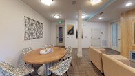 Located at 100 District Dr in Asheville, NC, this single-floor community amenity space offers a thoughtfully designed environment built for both relaxation and social engagement. The layout flows seamlessly through several distinct functional zones, each with its own character and purpose.

The entry corridor features crisp white walls adorned with contemporary abstract artwork in bold yellow and charcoal tones, setting a modern and welcoming tone. Patterned carpet tiles in neutral earth tones run throughout the space, adding texture and visual warmth underfoot.

The media lounge is a standout feature, anchored by a warm natural wood accent wall housing two large flat-screen televisions set within teal-trimmed built-in cabinetry with mesh-front storage below. Comfortable upholstered seating is arranged to face the screens, making it an ideal gathering spot for entertainment.

Adjacent to the media area, a lounge nook offers a more intimate seating arrangement, highlighted by a striking full-wall graphic wallcovering featuring a repeating vintage radio motif, lending a playful and creative energy to the space.

A dedicated dining area accommodates small group gatherings around a round wood-topped table with stylishly patterned accent chairs. Recessed and flush-mount ceiling lighting throughout ensures even, comfortable illumination across all areas. The overall ambiance is clean, contemporary, and inviting.