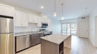 This thoughtfully designed one-bedroom, one-bathroom residence in Rosemount, Minnesota offers a functional and modern living experience. The open-concept kitchen is a standout feature, showcasing a two-tone cabinet design with crisp white upper cabinets and rich dark lower cabinets, granite countertops, a central island with built-in shelving, pendant lighting, and a full suite of stainless steel appliances including a refrigerator, range, microwave, and dishwasher. Light hardwood-style flooring flows throughout the kitchen and living areas, which benefit from natural light through sliding glass doors that open to an outdoor balcony view. The bedroom is a comfortable, carpeted retreat with a large window offering views of the surrounding area, a dedicated closet, and direct access to the full bathroom. The bathroom is finished with clean white cabinetry topped with granite, light tile flooring, and a tub-shower combination. A stacked in-unit washer and dryer, visible in the dollhouse view, adds everyday convenience. The overall aesthetic is clean, contemporary, and move-in ready.