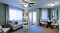 This single-floor space offers a comfortable and stylish living environment designed with both function and aesthetics in mind. The main living area immediately draws attention with its warm hardwood-style flooring, soft sage-green walls, and an abundance of natural light streaming through large windows dressed in sheer white curtains with decorative patterned accents. A ceiling fan with integrated lighting anchors the room, providing both airflow and ambient illumination. The centerpiece of the living area is a set of elegant French doors that open to an outdoor view, seamlessly connecting the interior to the outside environment and enhancing the sense of openness. The living space is thoughtfully arranged, featuring a plush sofa accented with blue throw pillows, decorative white garden stools serving as a coffee table, and a patterned area rug that ties the room together. A dining area is nestled nearby, with a compact table and upholstered chairs, making efficient use of the available space. The overall ambiance is bright, relaxed, and inviting, with a coastal-inspired color palette of whites, blues, and greens that creates a calm, welcoming atmosphere throughout.
