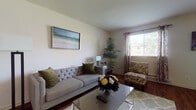 This single-floor residence offers a thoughtfully designed layout that balances comfort and functionality across its key living spaces. The living room serves as the heart of the home, featuring rich hardwood flooring, a tufted gray sofa, a glass-top coffee table, and a large window dressed with decorative curtains that invite natural light. A framed seascape painting and an indoor palm plant add to the room's relaxed, curated aesthetic. A wall-mounted flat-screen TV and a floating wooden shelf provide additional utility in the space.

The adjacent dining area continues the hardwood flooring and features a round table surrounded by teal upholstered chairs, pendant lighting overhead, and its own window for natural brightness.

The kitchen is well-equipped with rich wood cabinetry, granite-style countertops, stone-look tile flooring, and a full suite of stainless steel appliances including a refrigerator, range, microwave, and dishwasher.

Both bedrooms are carpeted and comfortably sized, each with a window allowing natural light. The primary bedroom includes a tufted upholstered headboard, while the second bedroom features a decorative metal bed frame.

Two bathrooms serve the residence — one with a walk-in glass-door shower and a dual-sink vanity with rich wood cabinetry, and a second with a tub-shower combination and matching tile flooring.