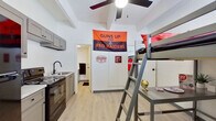 This compact studio is a single-floor unit thoughtfully designed to maximize functionality within a modest footprint. The space is organized into three distinct zones: a living/sleeping area, a kitchenette, and a private bathroom.

The main living area features light hardwood-style flooring, white walls, and a ceiling fan that contributes to a bright, airy feel. A loft-style bed frame with an integrated ladder and study desk beneath makes excellent use of vertical space, offering both a sleeping area and a dedicated workspace without compromising floor area.

The kitchenette is efficiently outfitted with gray shaker-style cabinetry — both upper and lower — complemented by light countertops. Appliances include a black electric range and a microwave, along with a single-basin sink with a modern matte black faucet. This kitchen area provides all the essential amenities for everyday meal preparation.

The private bathroom, accessed through a separate door, features a toilet and compact layout with tiled flooring. Exposed painted cinder block walls in a secondary area of the unit add an industrial character to the overall aesthetic.

A window allows natural light into the space, and the unit includes an entry door painted in a bold black finish. Overall, the studio presents a practical and functional layout well-suited for a single occupant seeking a self-contained living environment.